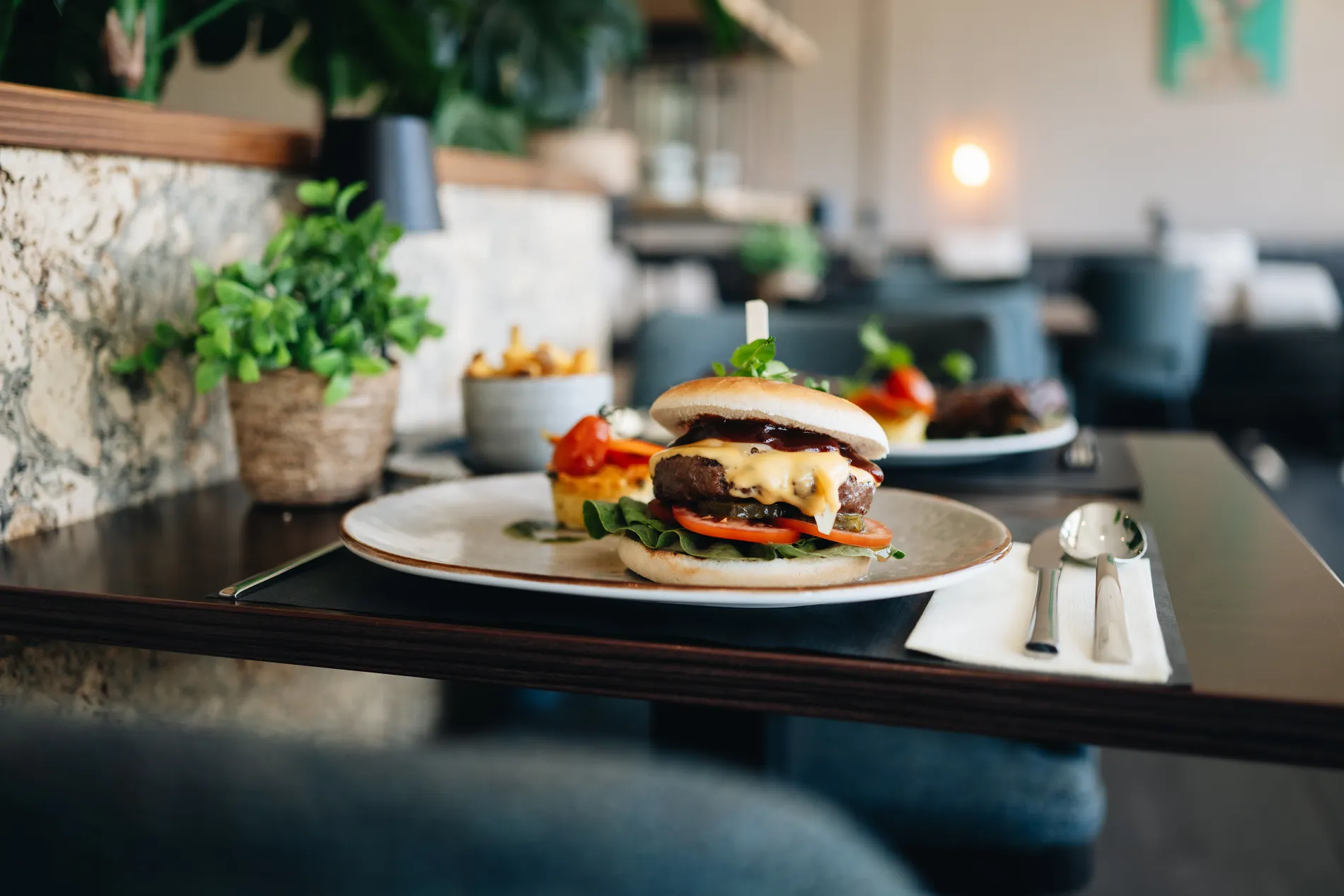 Een sappige hamburger met gesmolten kaas, tomaat, augurk en sla op een bord in het sfeervolle interieur van het restaurant, geserveerd met frites op de achtergrond.